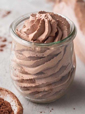 A small jar filled with chocolate whipped cream. A piping bag filled with whipped cream and a small bowl of cocoa powder rest nearby.