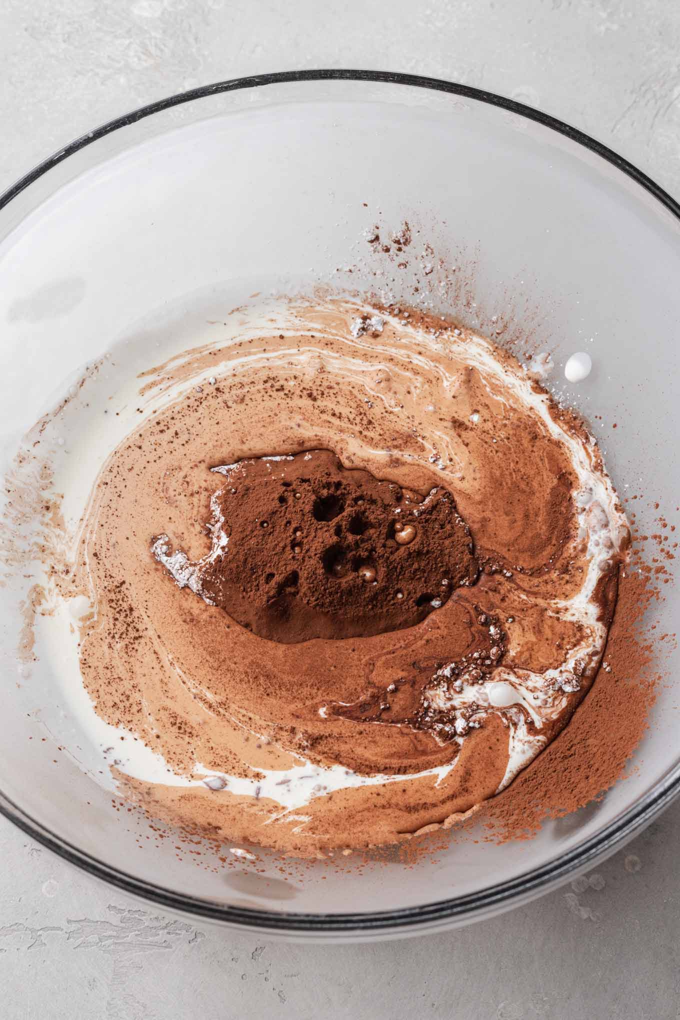 A glass mixing bowl with heavy cream, powdered sugar, cocoa powder, and vanilla extract.