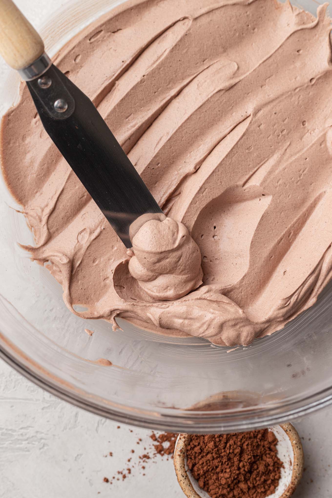 A glass mixing bowl filled with the finished chocolate whipped cream. An angled spatula is resting inside of the bowl.