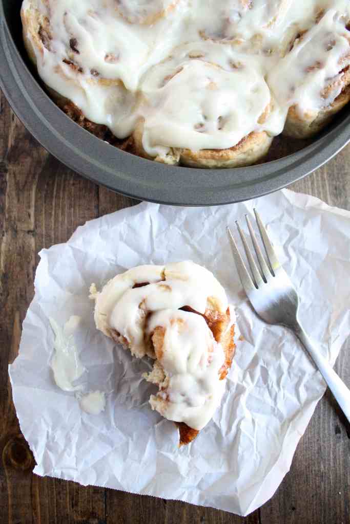 Overhead view of 1 hour cinnamon rolls in a round pan. A half-eaten cinnamon roll and a fork rest nearby on a piece of paper. 