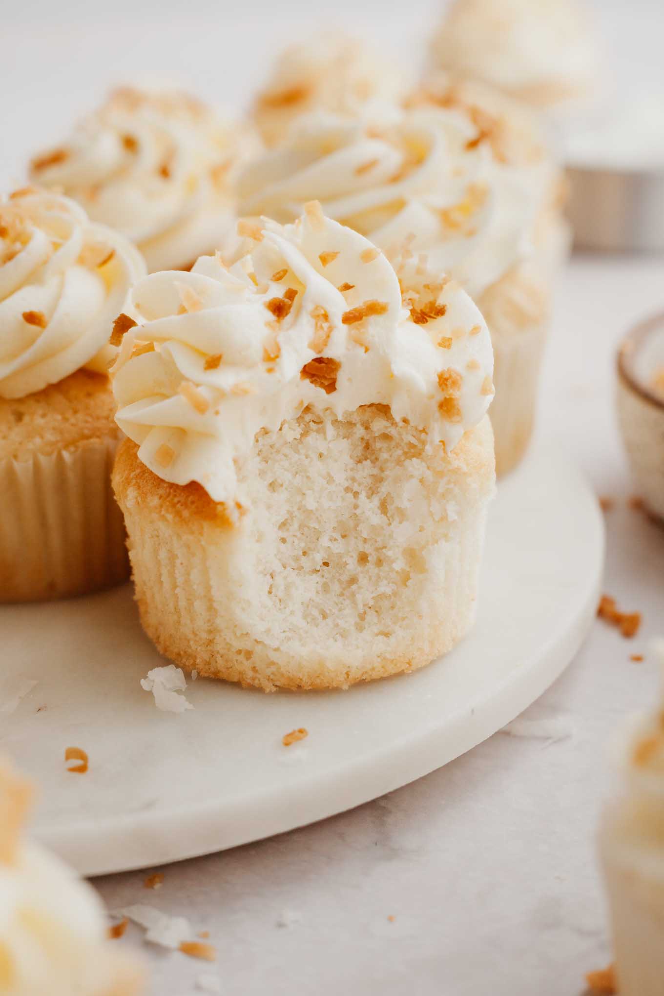 A coconut cupcake with a bite missing, with additional cupcakes on the plate behind it. 