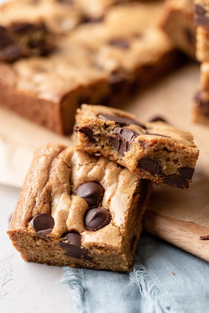 Two chocolate chip cookie bars resting on each other. The top top bar has a bite missing. More bars rest in the background.