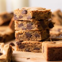 A stack of four chocolate chip cookie bars on a wood serving board. More cookie bars surround the stack.