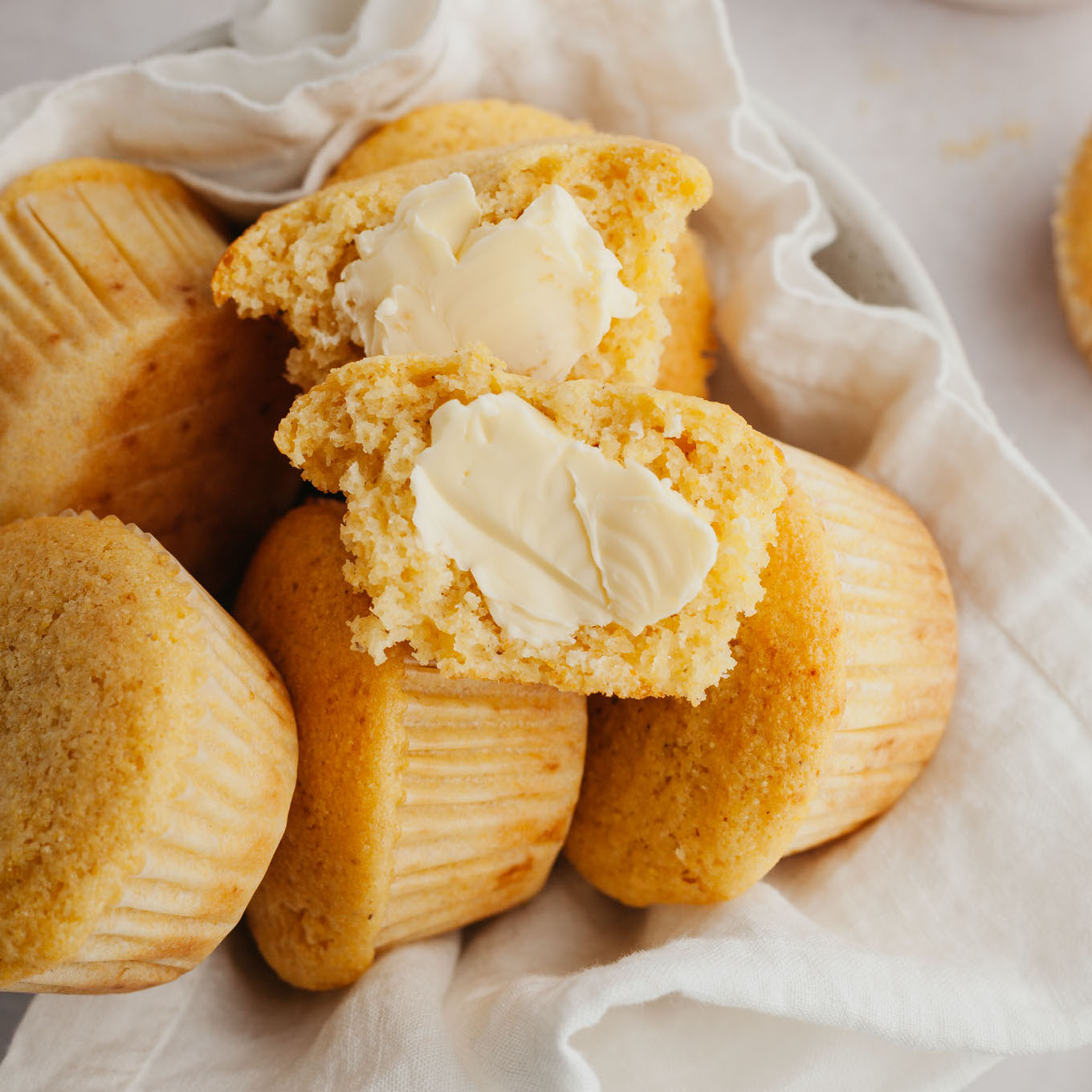 A pile of cornbread muffins in a towel-lined serving bowl. The top muffin has been split in half and slathered with butter. 