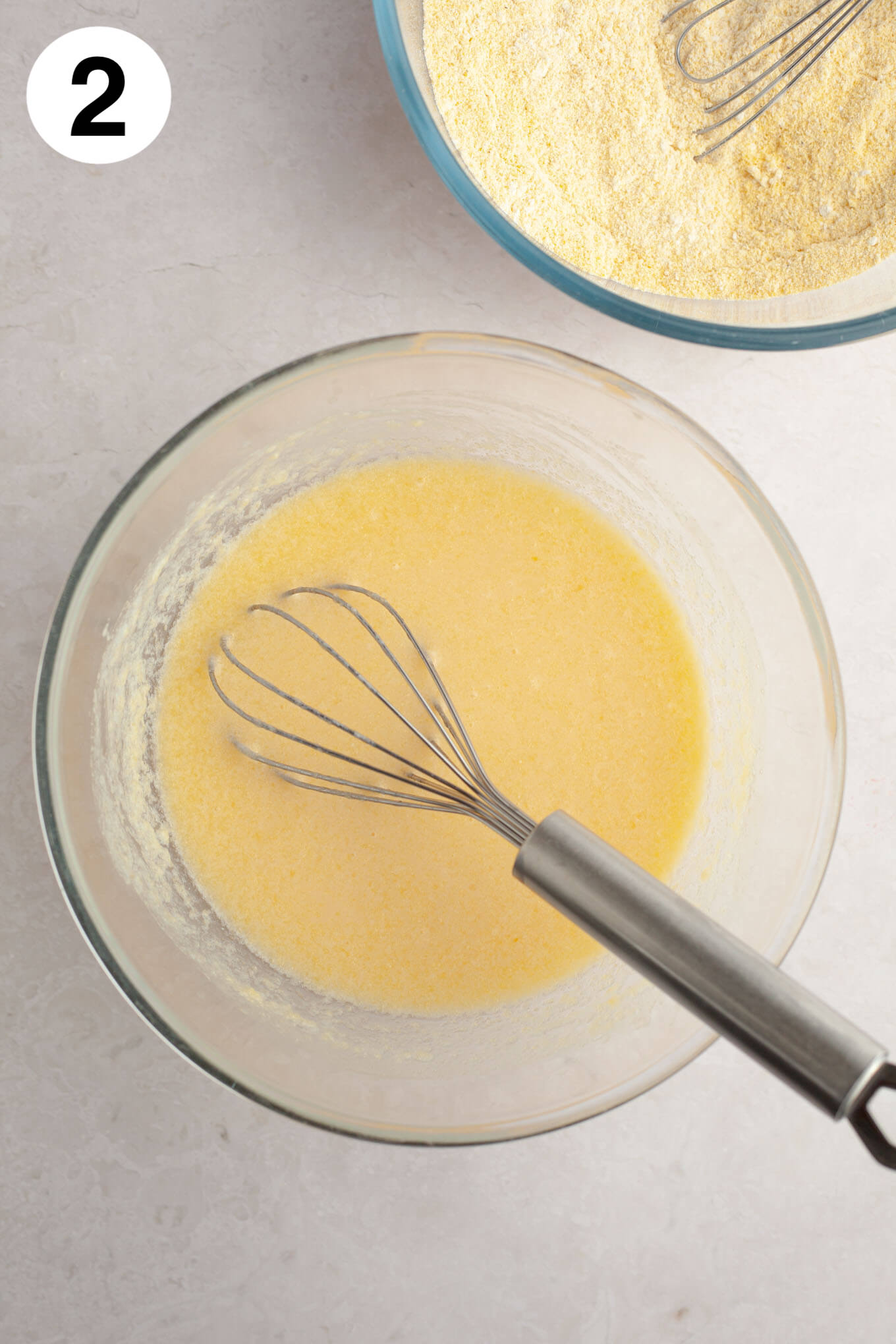 An overhead view of the wet ingredients for cornbread muffins in a glass mixing bowl with a wire whisk. 