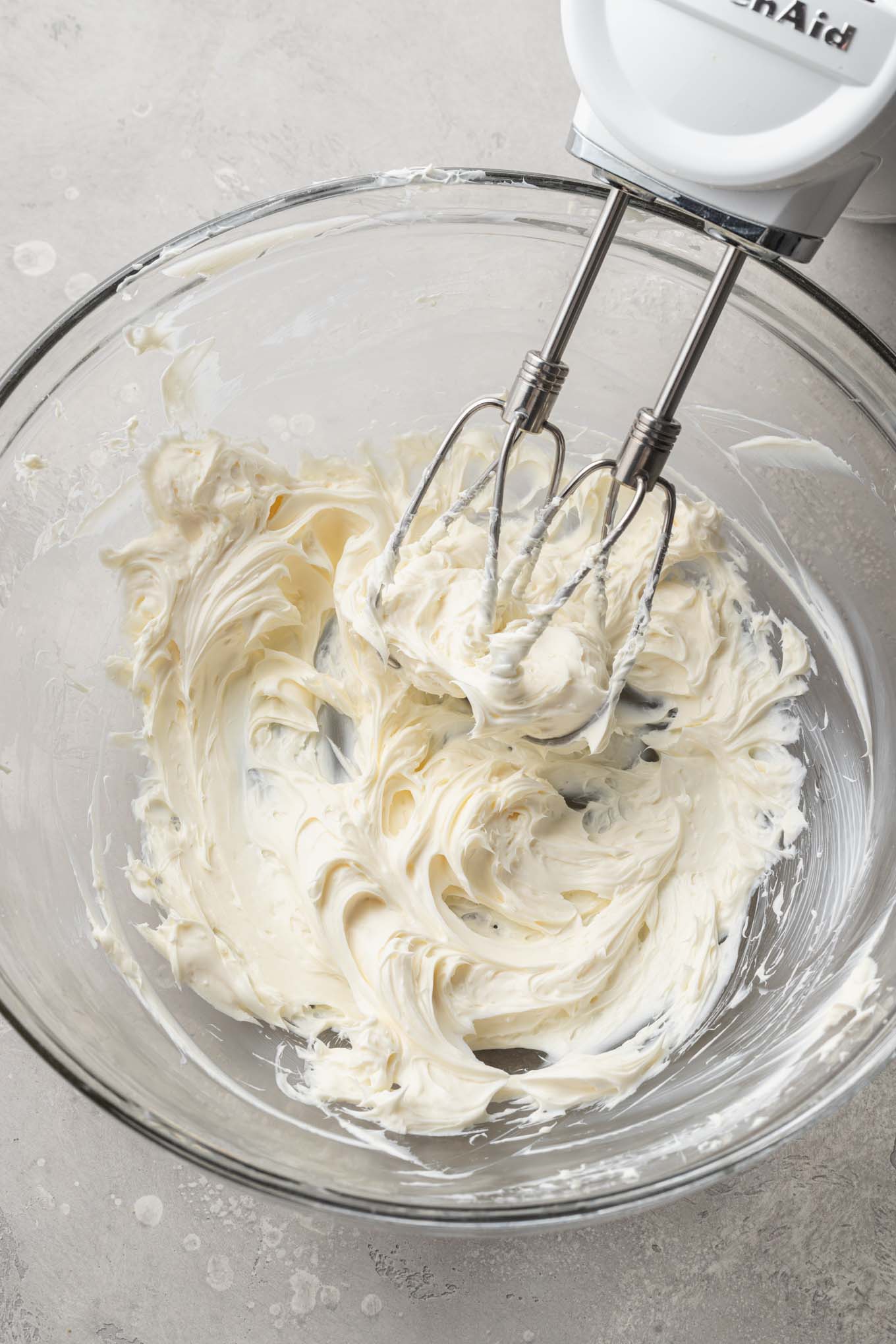 An overhead view of beaten cream cheese in a glass mixing bowl, with a handheld mixer. 