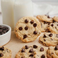 Several cream cheese chocolate chip cookies on a piece of brown parchment paper. One of the cookies is leaning against a glass of milk.