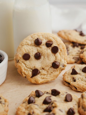 Several cream cheese chocolate chip cookies on a piece of brown parchment paper. One of the cookies is leaning against a glass of milk.