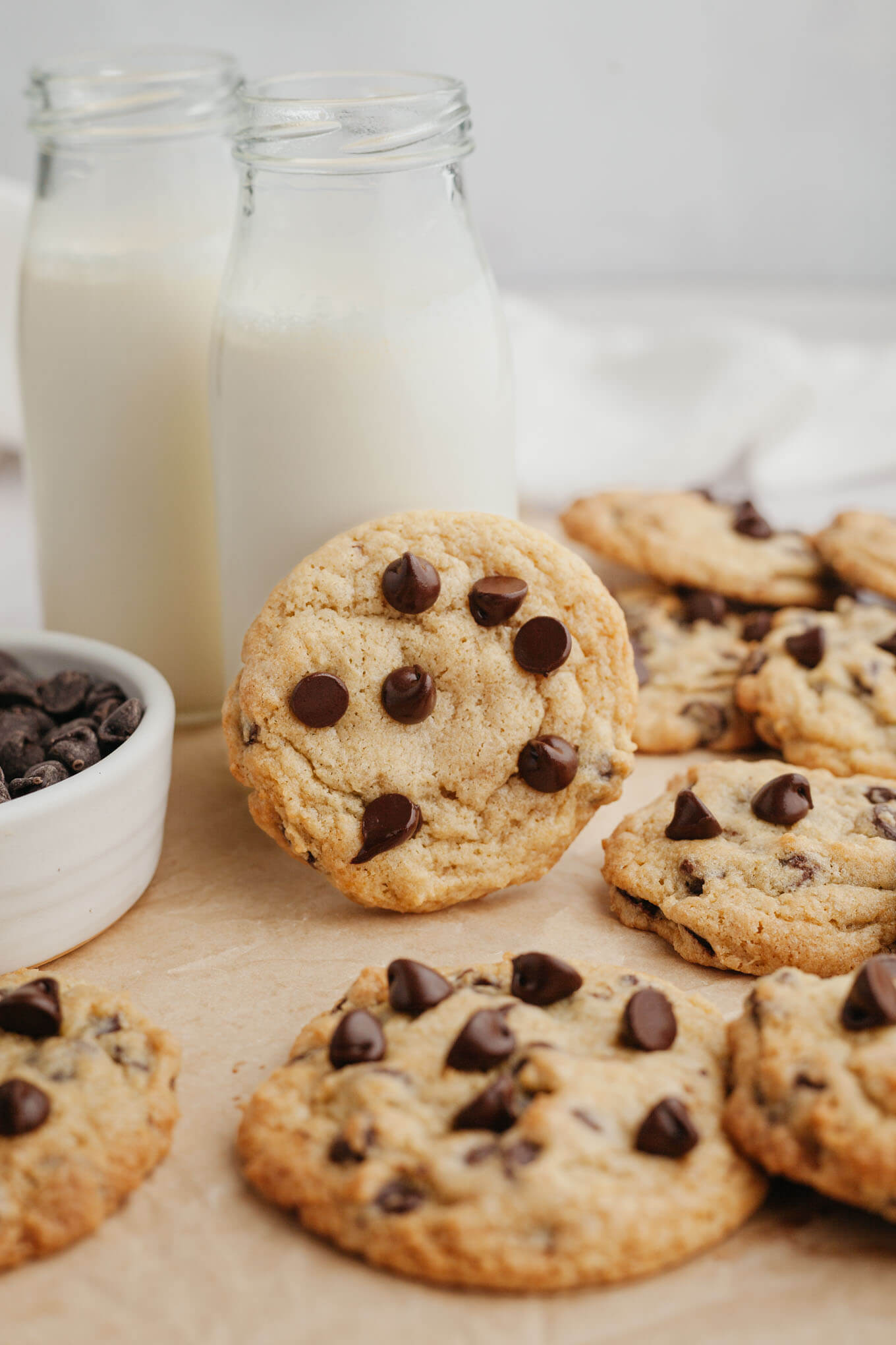 Two glass milk jugs surrounded by cream cheese chocolate chip cookies. One cookie is leaning against a milk jug. 