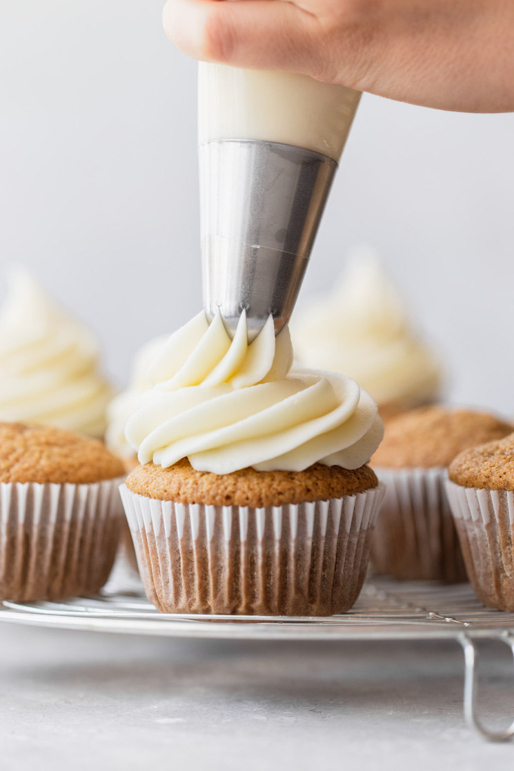 A group of cupcakes on a round cooling rack being topped with cream cheese frosting.