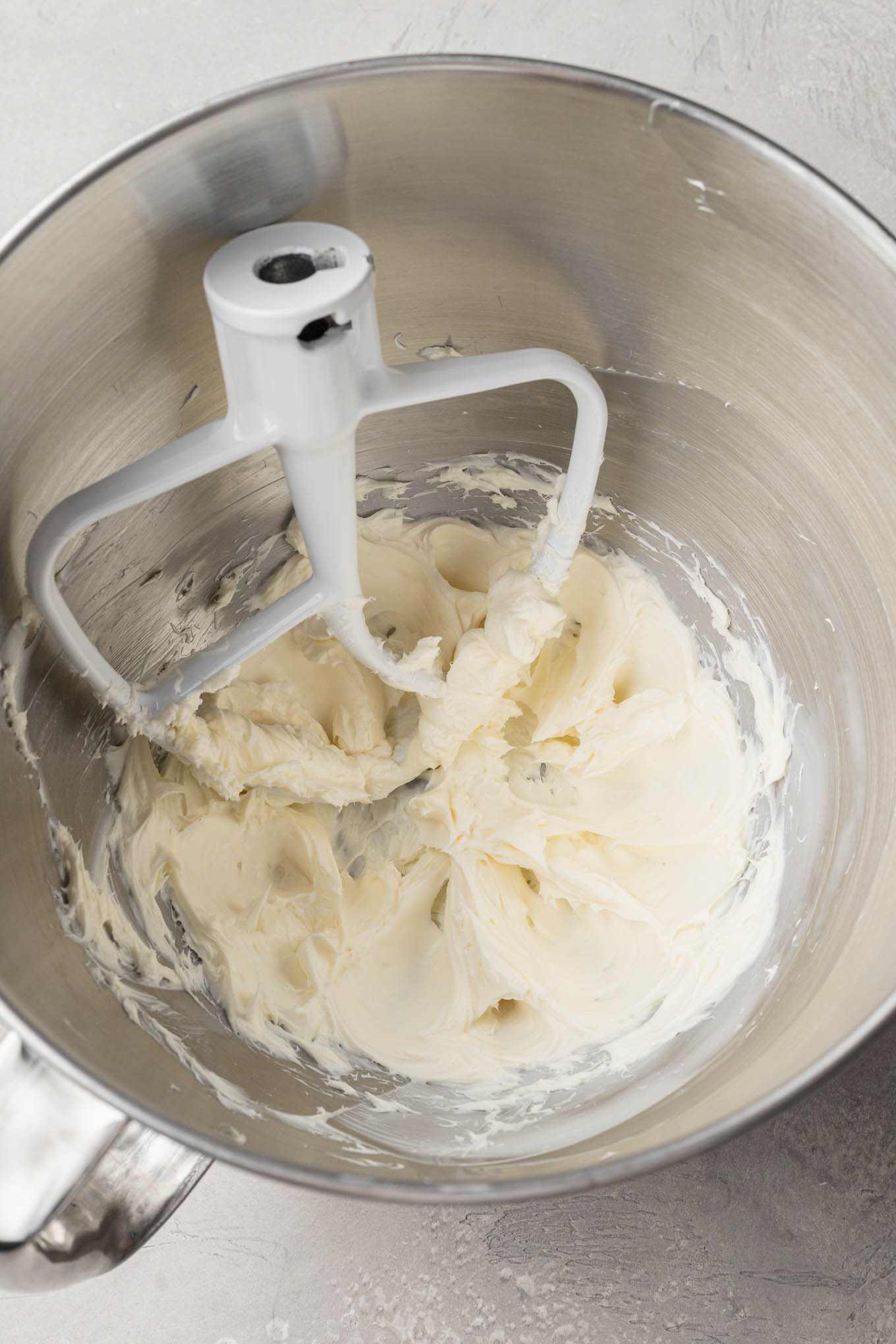 An overhead view of beaten cream cheese in a mixing bowl, with a paddle attachment. 