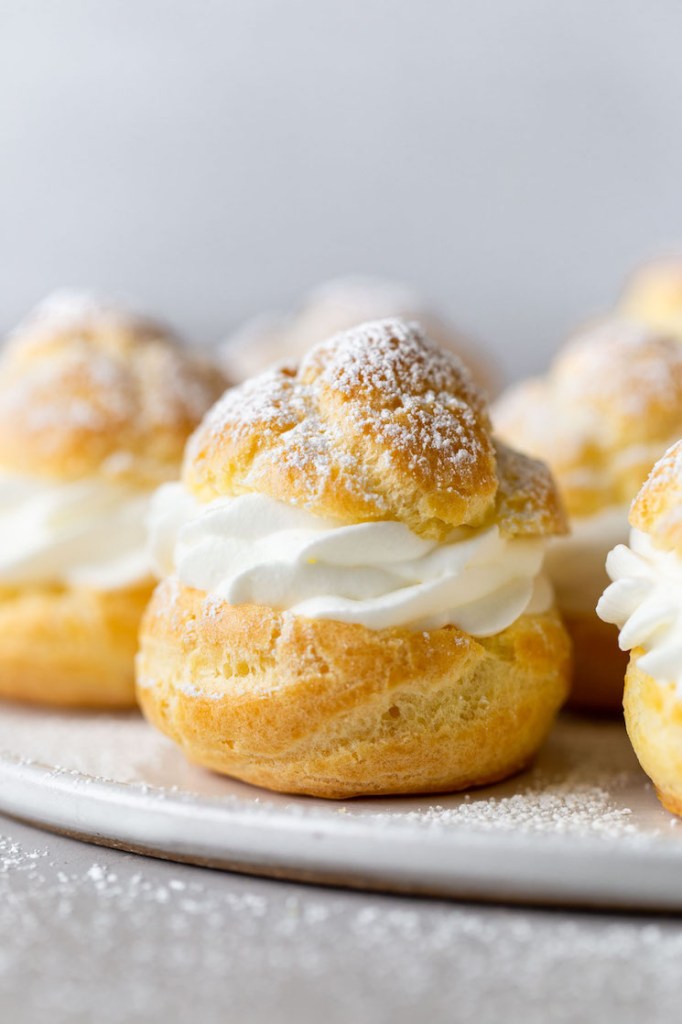 Side view of homemade cream puffs filled with whipped cream on a white plate. 