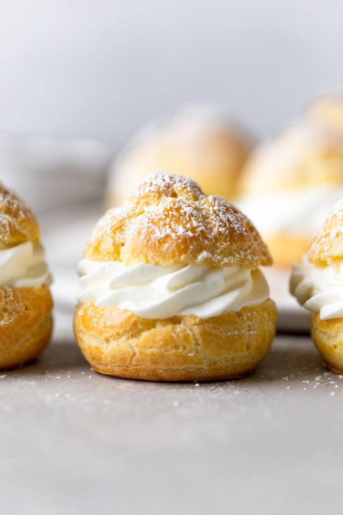 Side view of filled cream puffs lined up on a countertop. More cream puffs rest on a plate in the background.