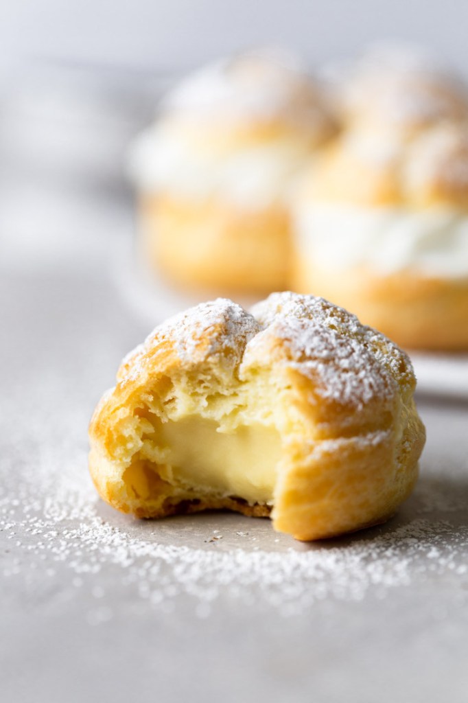 A cream puff with a bite missing. More filled cream puffs are on a plate in the background. 