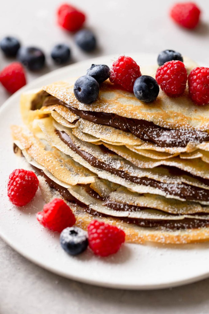 A stack of homemade crepes oozing Nutella and topped with fresh berries. More berries are scattered in the background. 