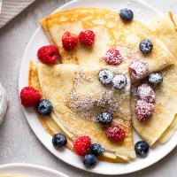 Overhead view of three homemade crepes topped with fresh berries and dusted with powdered sugar on a white plate.