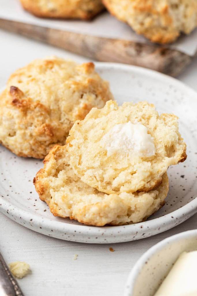 Two drop biscuits on a white plate. One biscuit has been cut in half and topped with butter. 