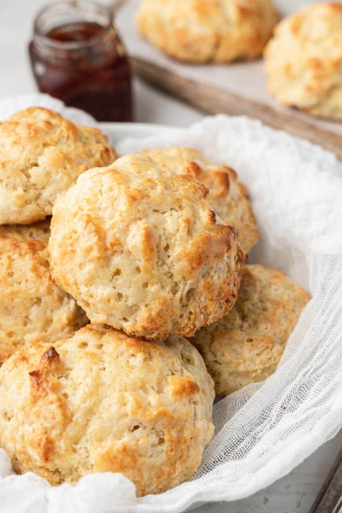 A pile of homemade drop biscuits in a bowl, with a jam jar in the background. 