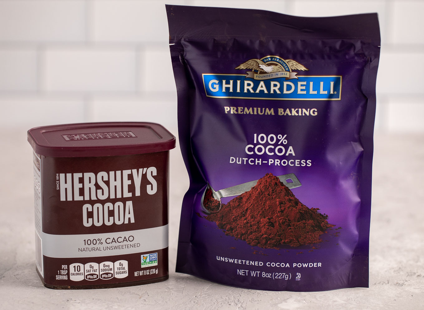 A container of Hershey's unsweetened cocoa powder and a package of Ghirardelli Dutch process cocoa powder sitting next to each other on a gray surface.