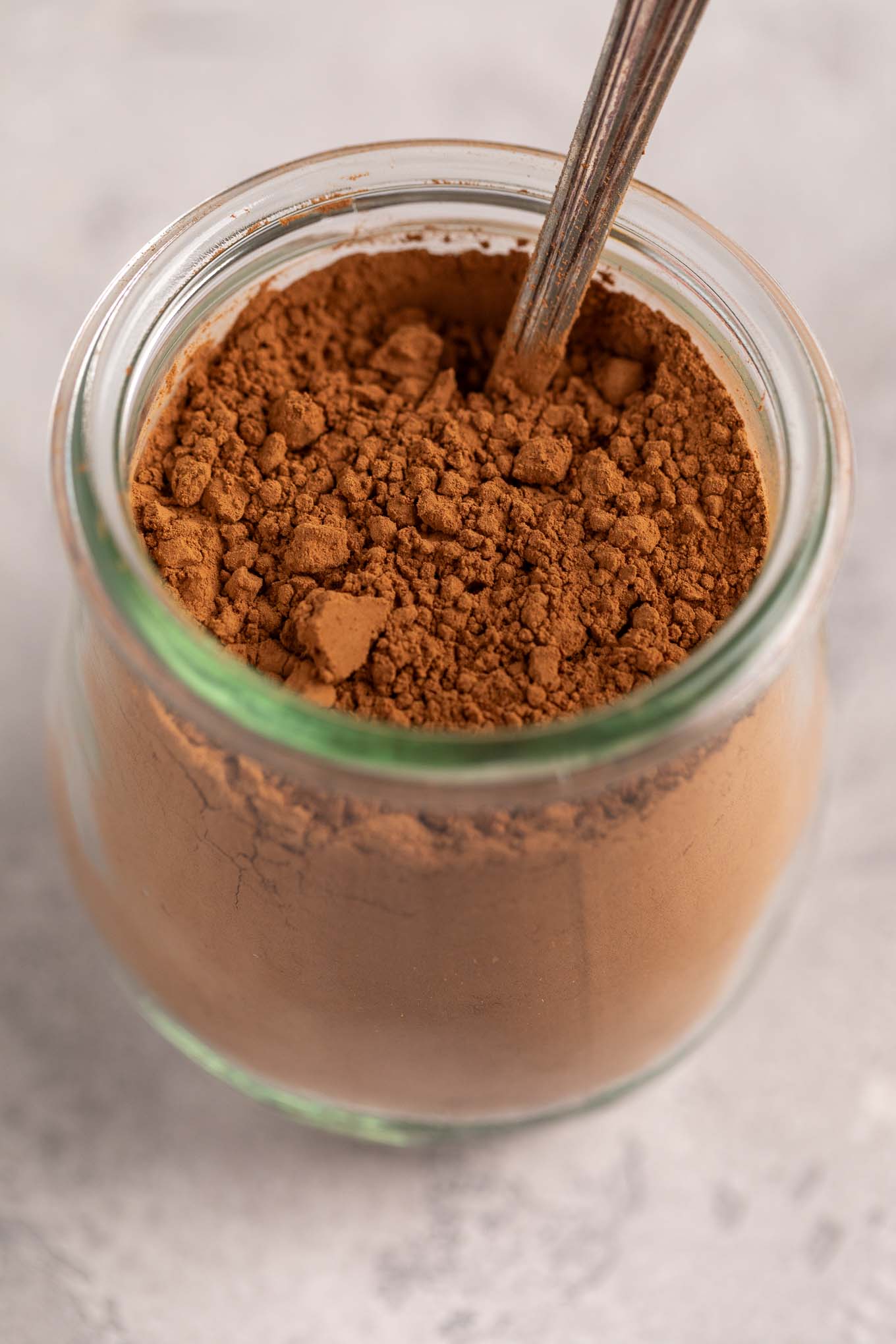 A small glass jar filled with cocoa powder. A spoon is resting inside of the jar.