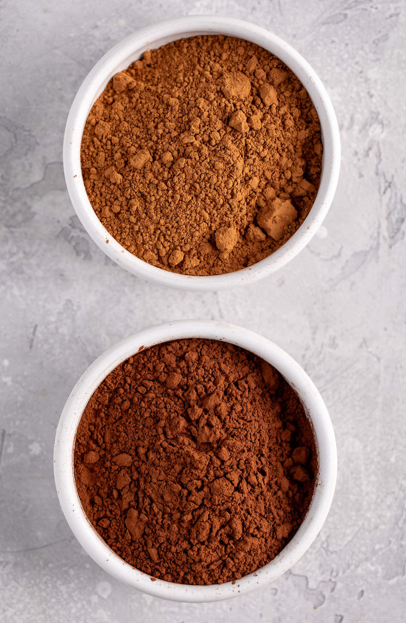 An overhead image of a white bowl filled with natural cocoa powder and another white bowl filled with Dutch process cocoa powder.