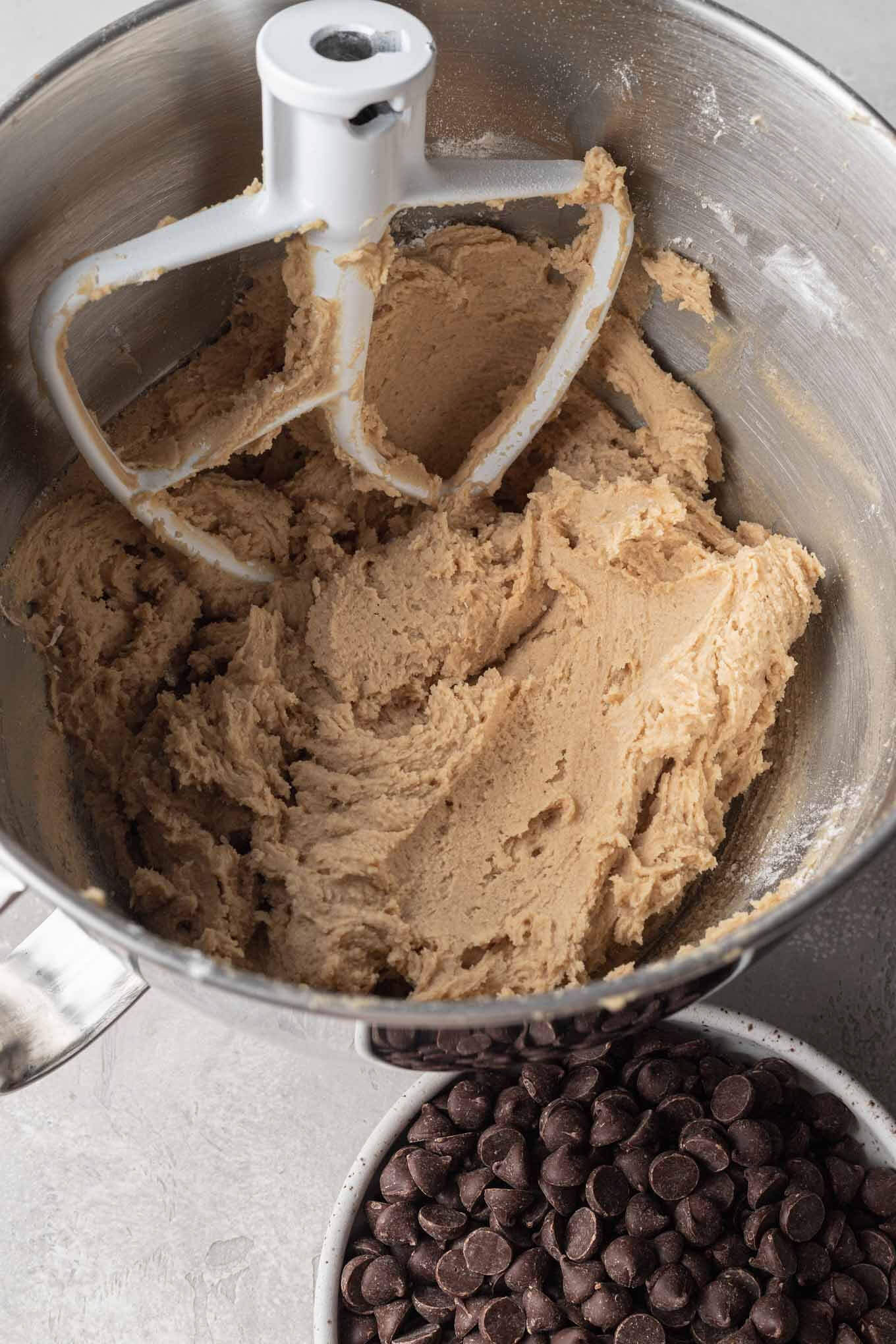 No-egg cookie dough in a metal mixing bowl with a paddle attachment. A bowl of chocolate chips rest on the side. 