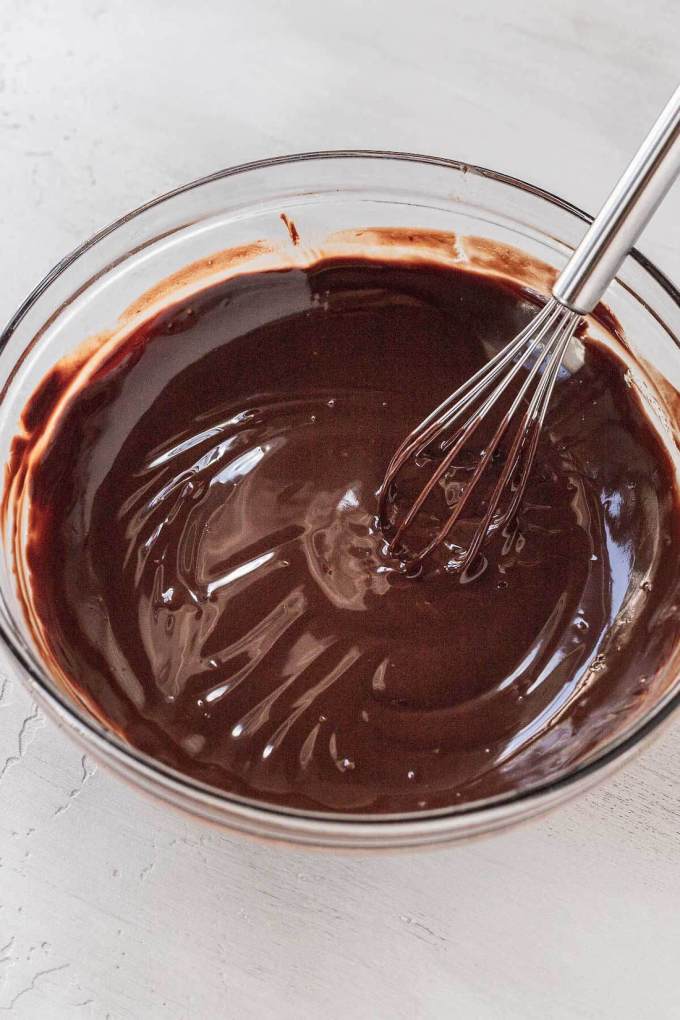 A glass mixing bowl full of melted chocolate. A whisk rests on the side.