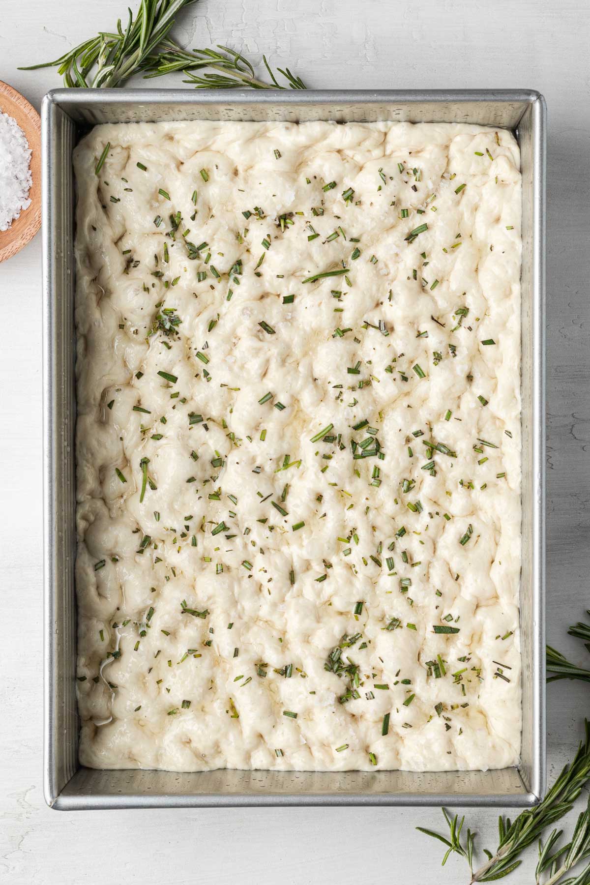 An overhead view of raw focaccia dough in a metal baking pan. 