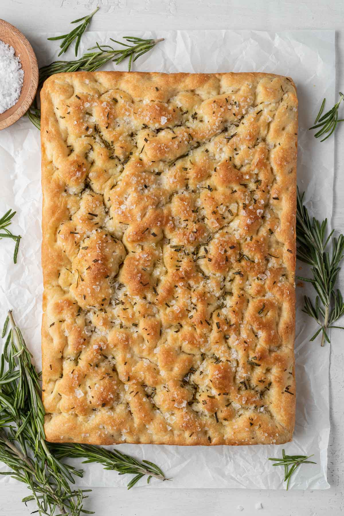 An overhead view of a loaf of baked focaccia, surrounded by fresh rosemary sprigs and a small bowl of sea salt. 