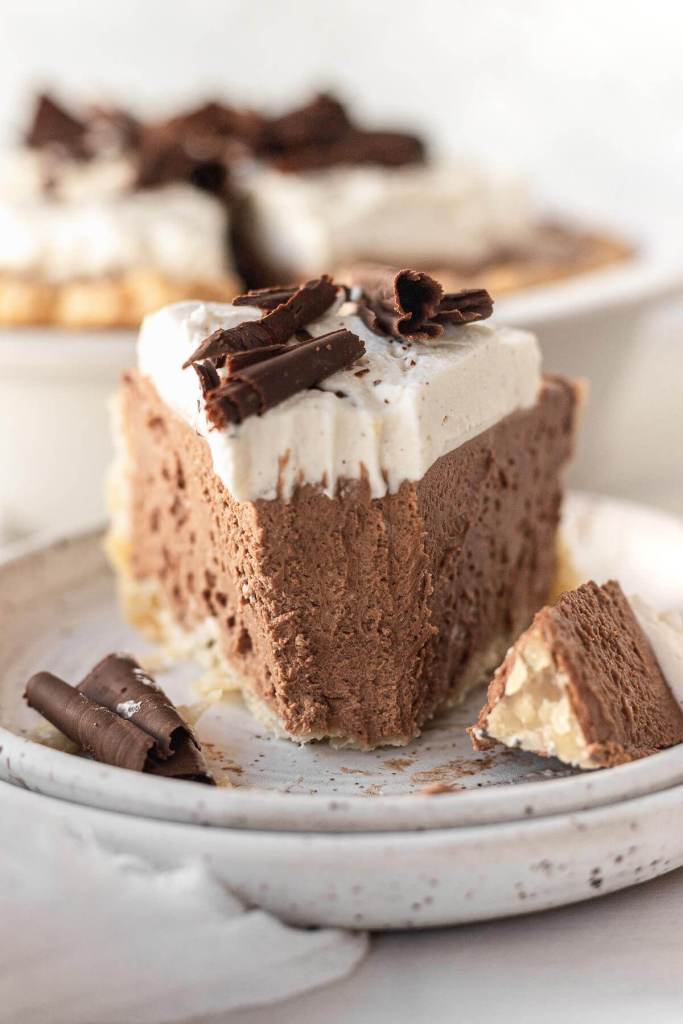 A front view of a slice of French silk pie on a white speckled plate, with a bite missing. The rest of the pie rests in the background. 