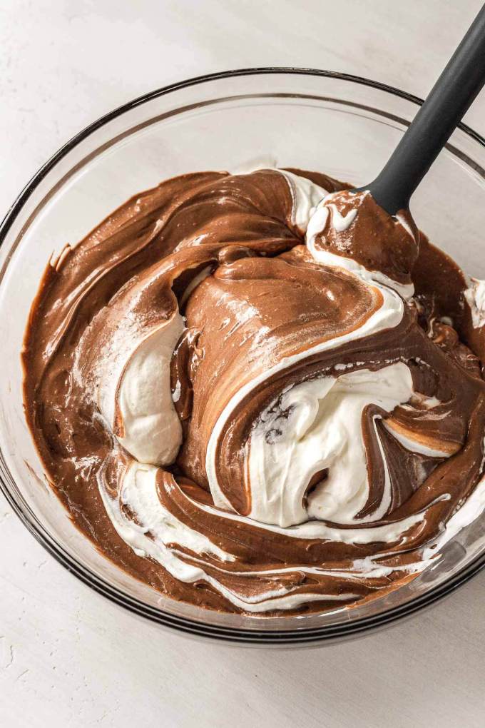 Whipped cream being mixed into a chocolate silk pie filling in a glass mixing bowl.