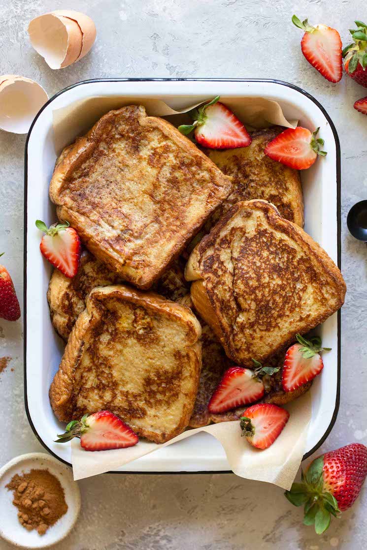 A baking pan filled with finished French toast surrounded by strawberries and spices.