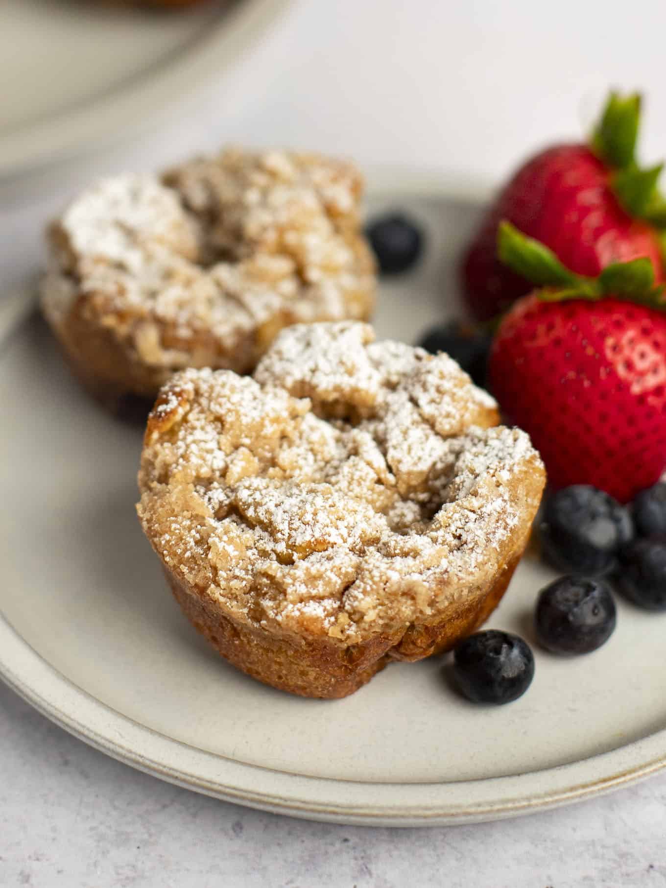 Two French toast muffins dusted with powdered sugar sitting on a plate next to fresh fruit.