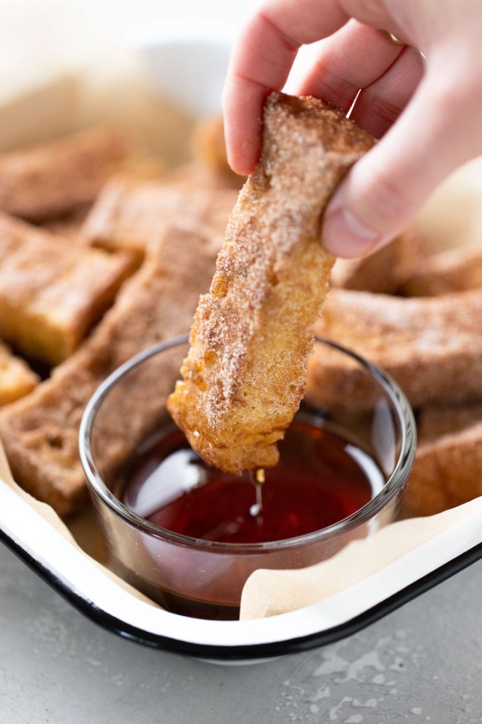 A baked french toast stick being dunked into a small dish of maple syrup. 