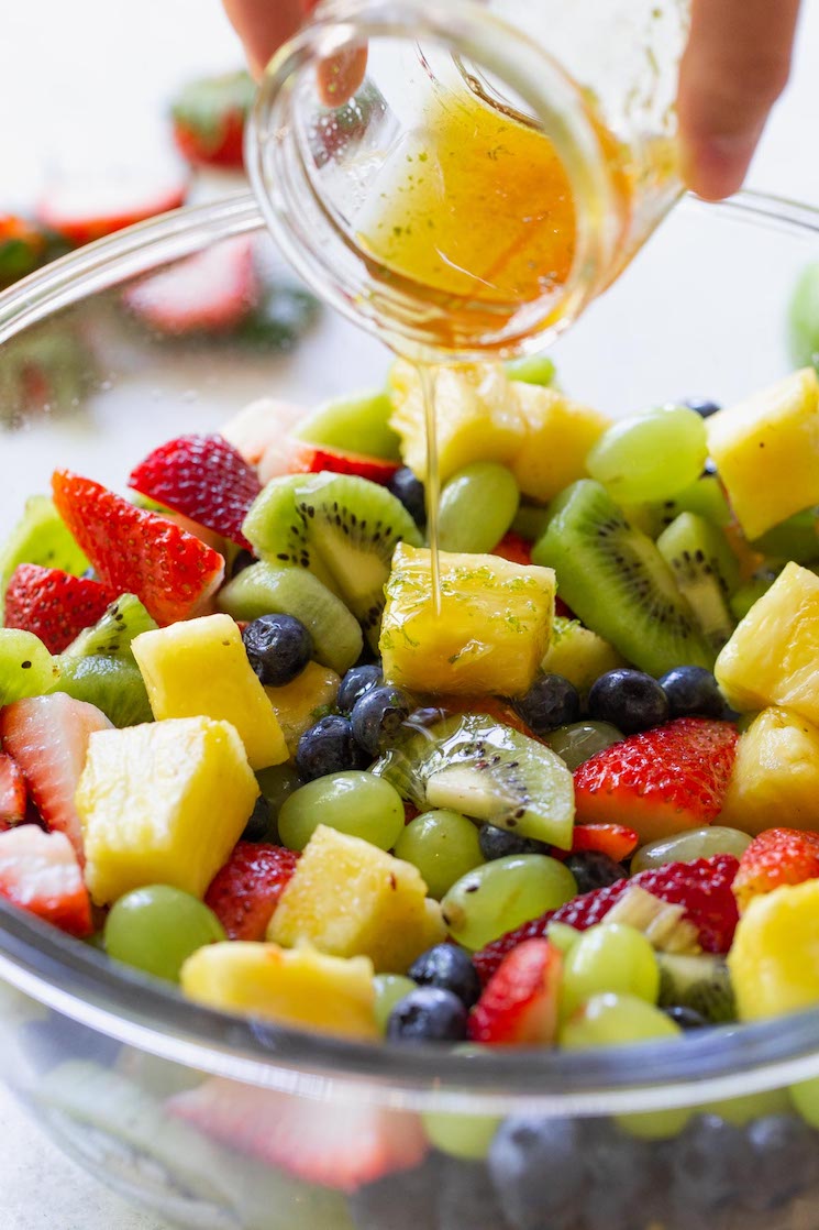 A glass bowl filled with fruit salad and the honey lime dressing being poured out of a glass jar over it. 