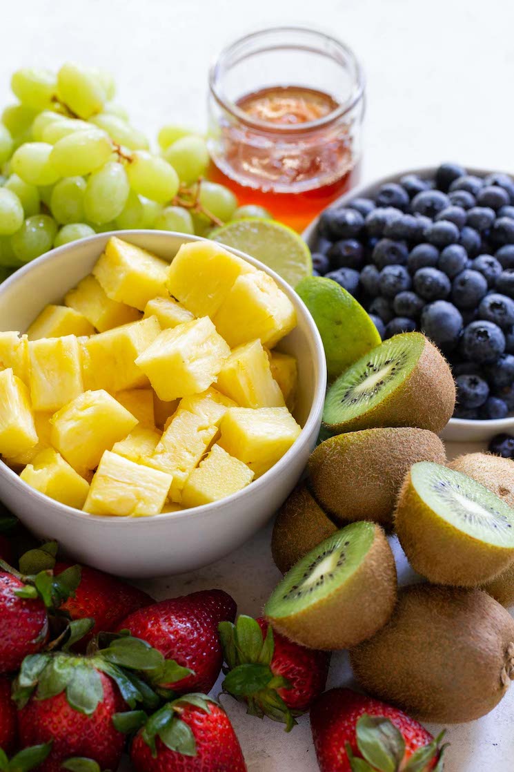 All of the fruit needed to make fruit salad prepared and laying on a rustic gray surface.