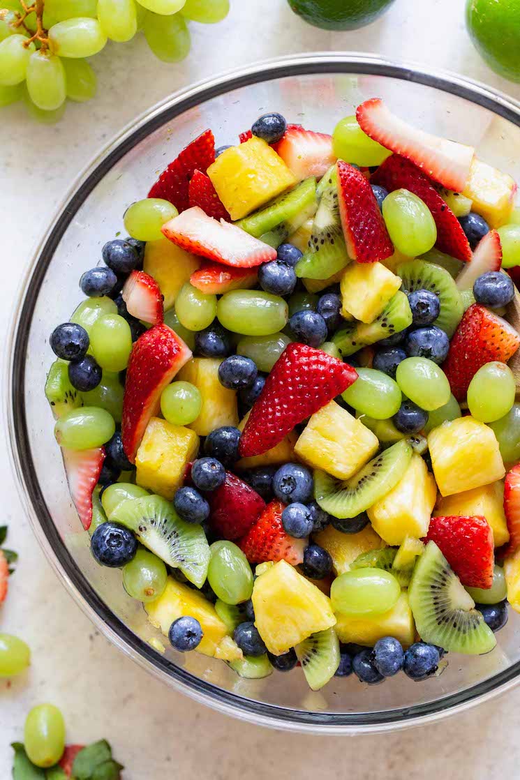 A glass bowl filled with fruit salad on a rustic gray surface with more fruit displayed around it.