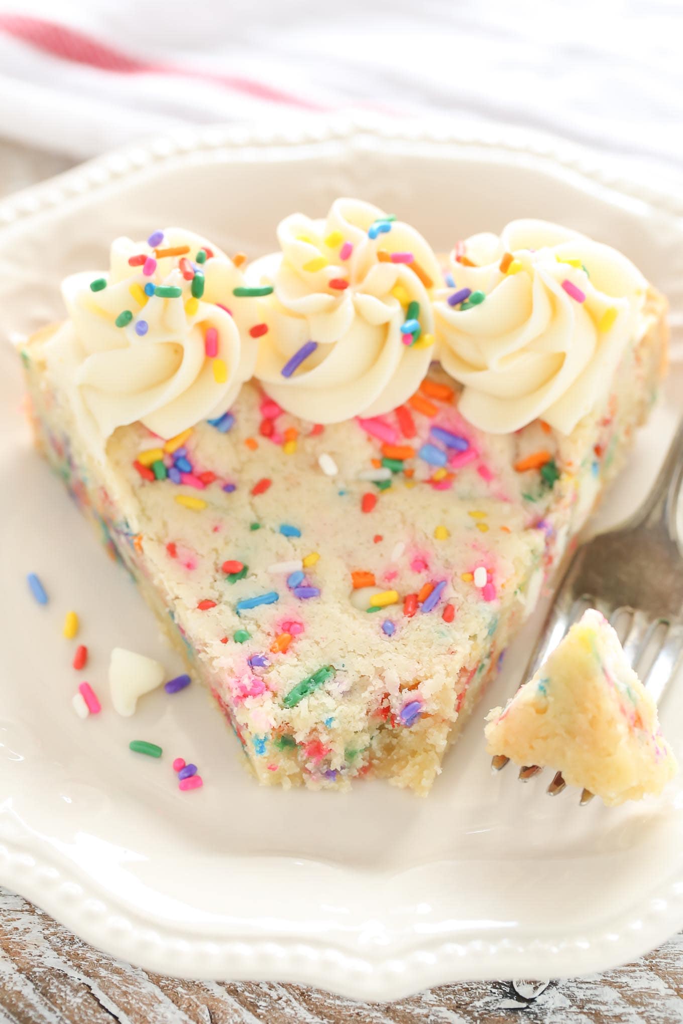 A slice of funfetti sugar cookie cake on a white plate with a fork. The fork has speared a bite of the cake. 