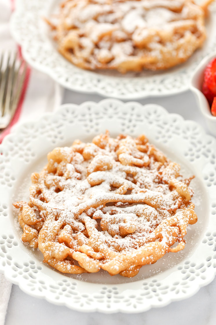 Two homemade funnel cakes topped with powdered sugar on decorative white plates and strawberries on the side. 