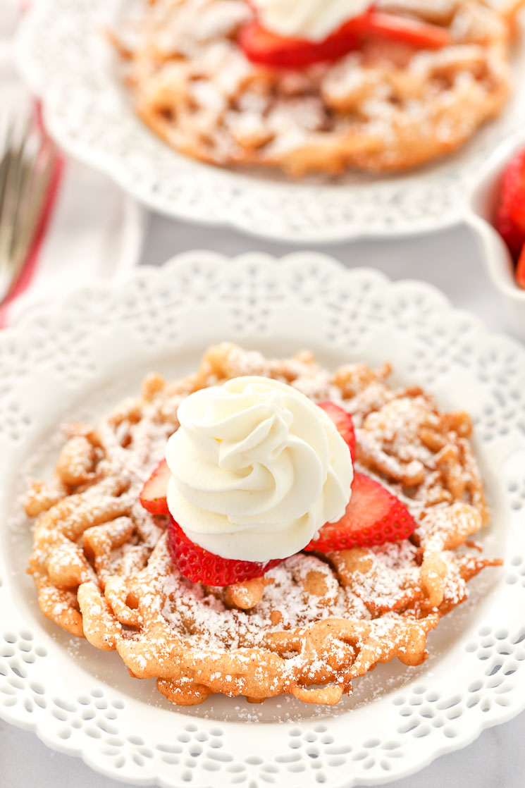 Two funnel cakes topped with powered sugar, strawberries, and homemake whipped cream sitting on decorative white plates. 