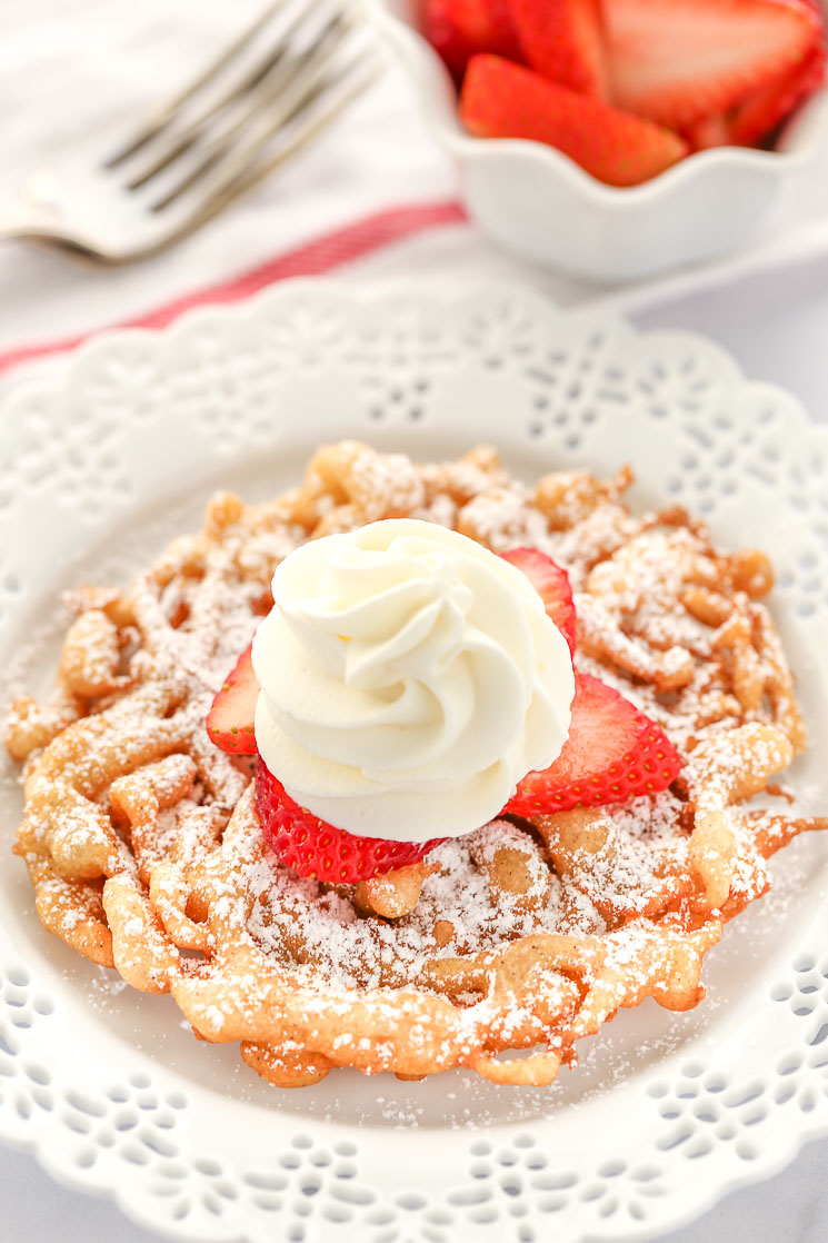 A homemade funnel cake topped with strawberries and homemade whipped cream sitting on a decorative white plate.