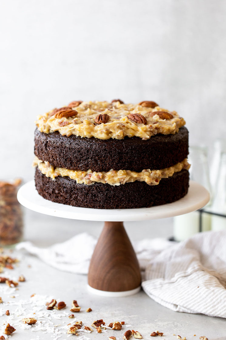 A layered German Chocolate Cake on a marble cake stand with chopped pecans and coconut around it.