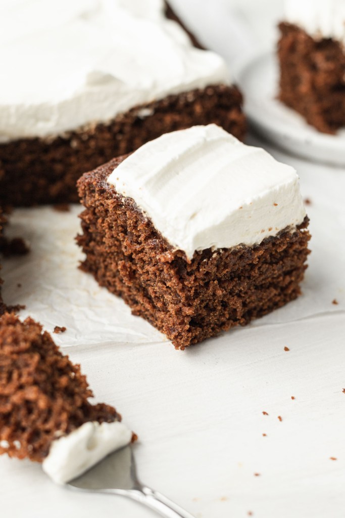 A slice of gingerbread cake topped with whipped cream. The rest of the cake is behind it. 