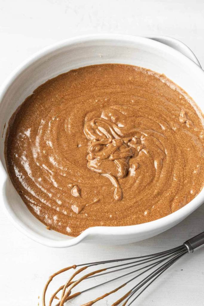 Gingerbread cake batter in a large white mixing bowl. A batter-covered whisk rests on the side. 