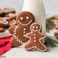 Two iced gingerbread cookies propped up a against a small jug of milk. More cookies rest on a plate in the background.