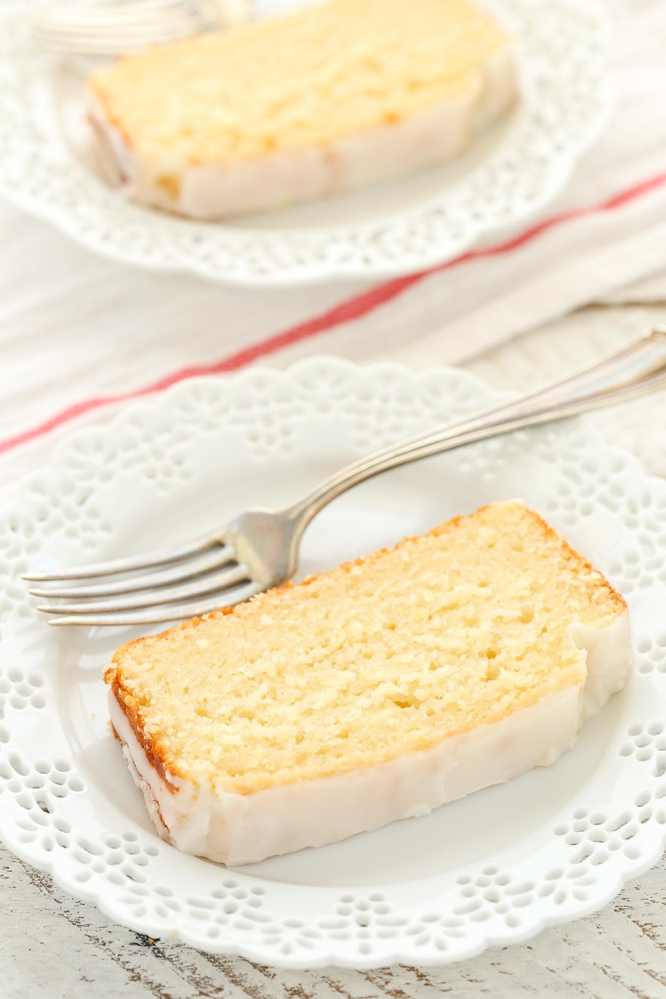 A slice of lemon bread on a white plate with a fork. Another plate rests in the background. 