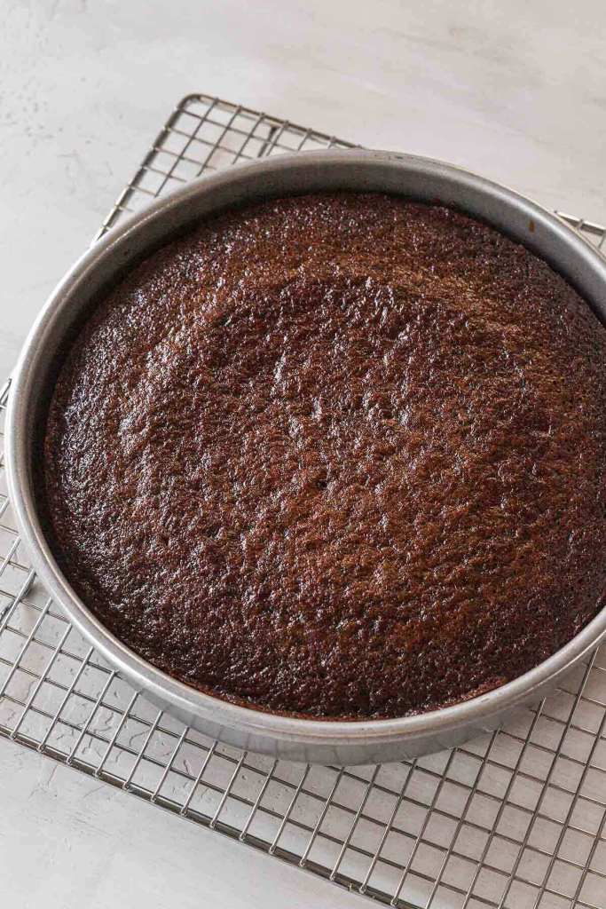 A baked chocolate cake cooling in its pan atop a wire rack. 