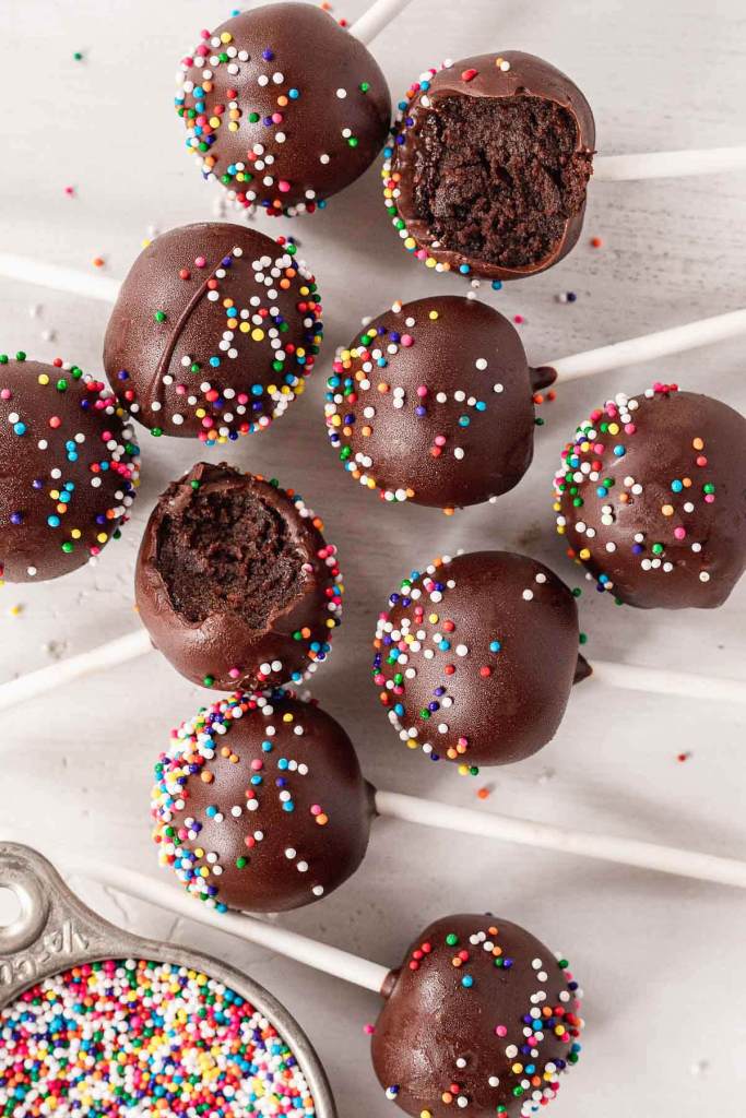 An overhead view of cake pops dipped in chocolate and covered in sprinkles. Two cake pops have bites missing. 