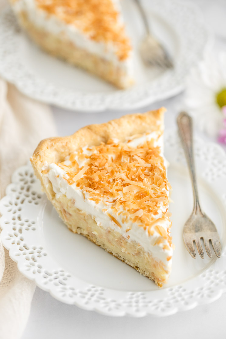 A slice of coconut cream pie on a decorative white plate with another slice in the background.