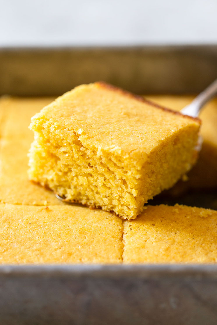 A finished pan of cornbread with one piece cut out and sitting on top showing the texture and golden color.